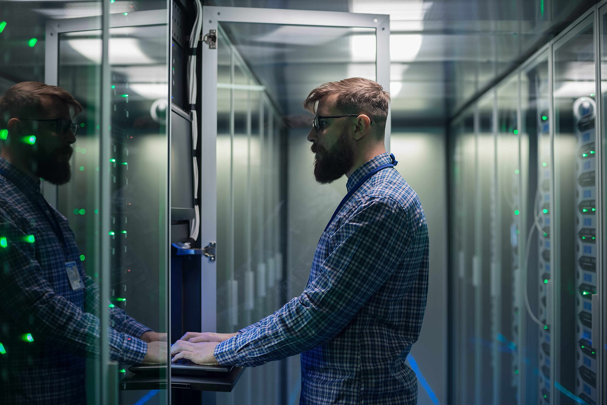 Ein Mann arbeitet an einem Computerterminal in einem Serverraum mit Glaswänden. Grüne Lichter blinken um ihn herum, während er sich auf die Bildschirme konzentriert.